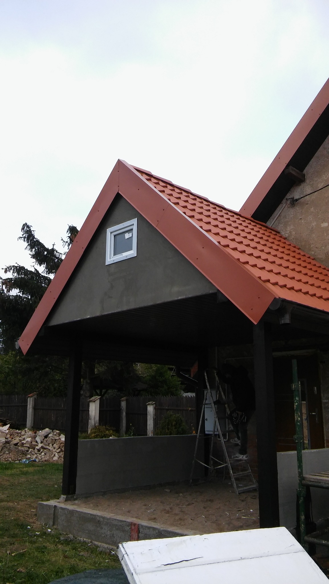 Zadaszenie tarasu z czerwoną dachówką, małym oknem i świeżo otynkowaną ścianą, widoczne drabina i materiały budowlane.