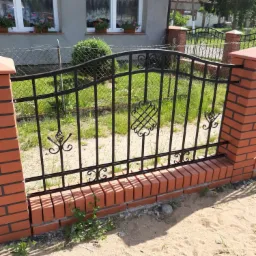 Fragment czarnego ogrodzenia z metalowymi zdobieniami, osadzonego na niskim murku z czerwonej cegły, z widocznym domem w tle i piaszczystym podłożem z pojedynczymi chwastami.
