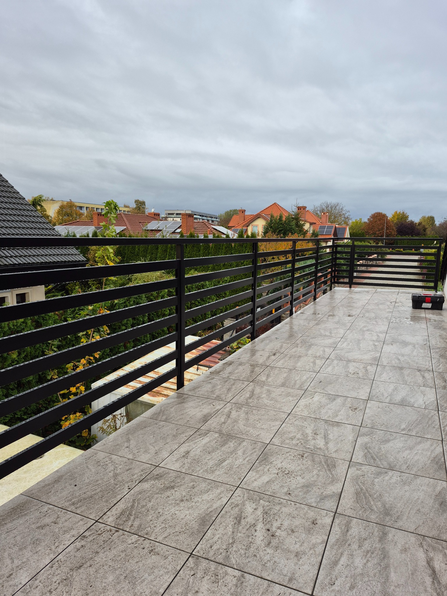 Nowoczesna, pozioma balustrada balkonowa z czarnych, płaskich elementów, na tle szarej, jesiennej scenerii i domów z czerwonymi dachami. Widoczna skrzynka narzędziowa.