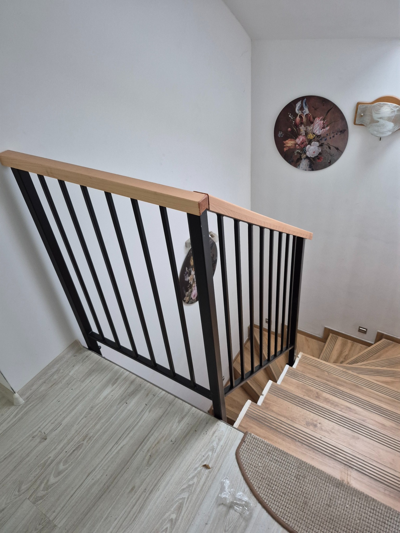 Nowoczesna balustrada schodowa z czarnymi, pionowymi prętami i drewnianym pochwytem, widok z góry na drewniane schody z nakładkami dywanowymi i dekoracją ścienną.