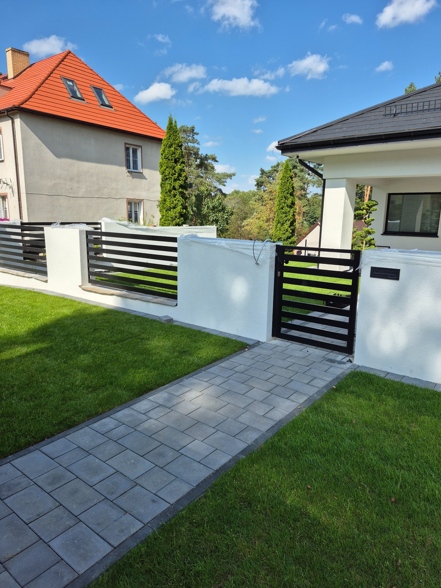 Nowoczesna brama wjazdowa z poziomymi, czarnymi przęsłami, wkomponowana w biały mur. Uwagę przykuwa starannie ułożony chodnik z kostki brukowej i soczysta zieleń trawnika.