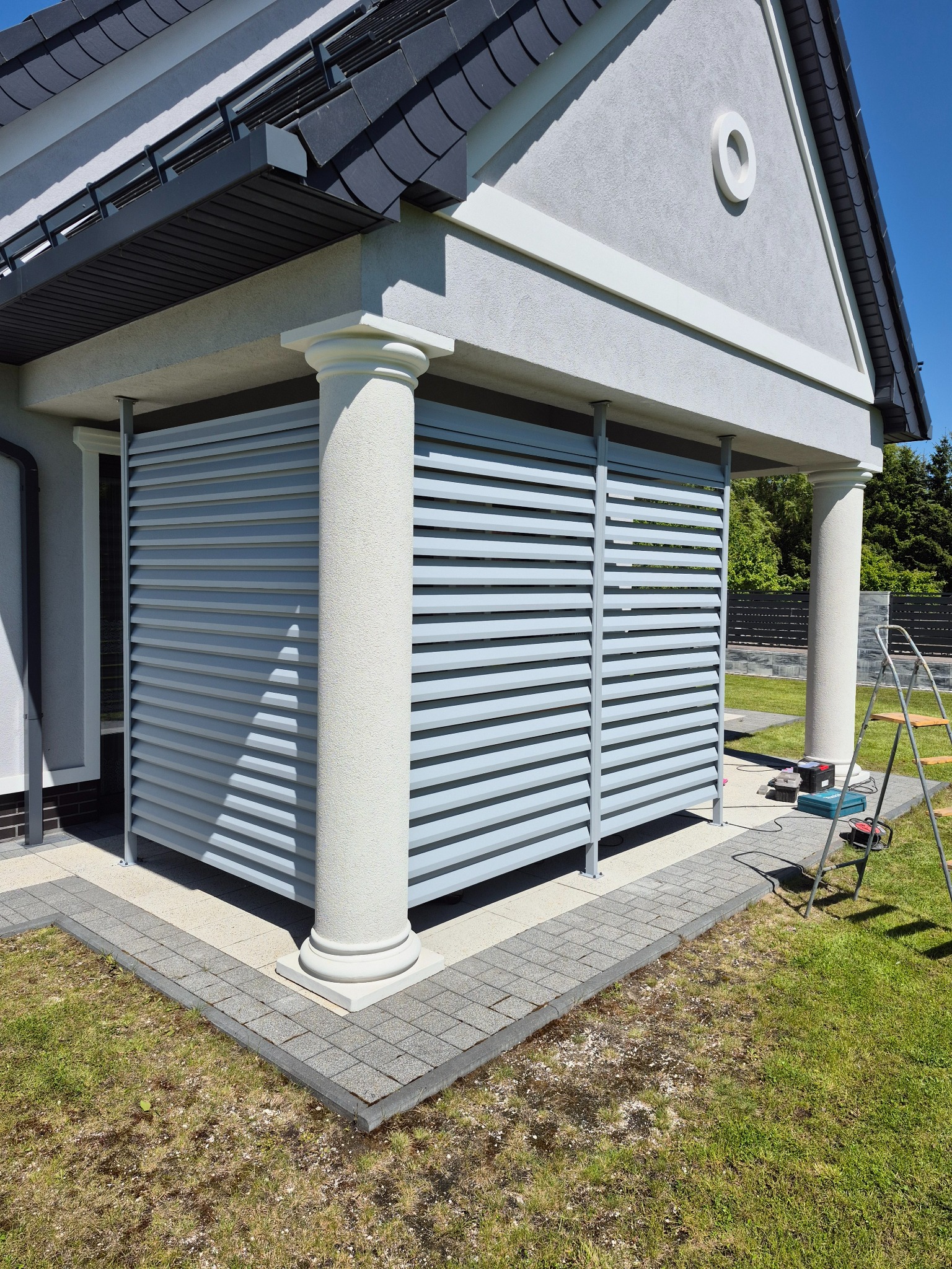 Nowoczesna, szara osłona tarasu z poziomych lameli, wsparta na kolumnach. Widoczne narzędzia i drabina sugerują montaż w toku. Jasne tło, trawnik.