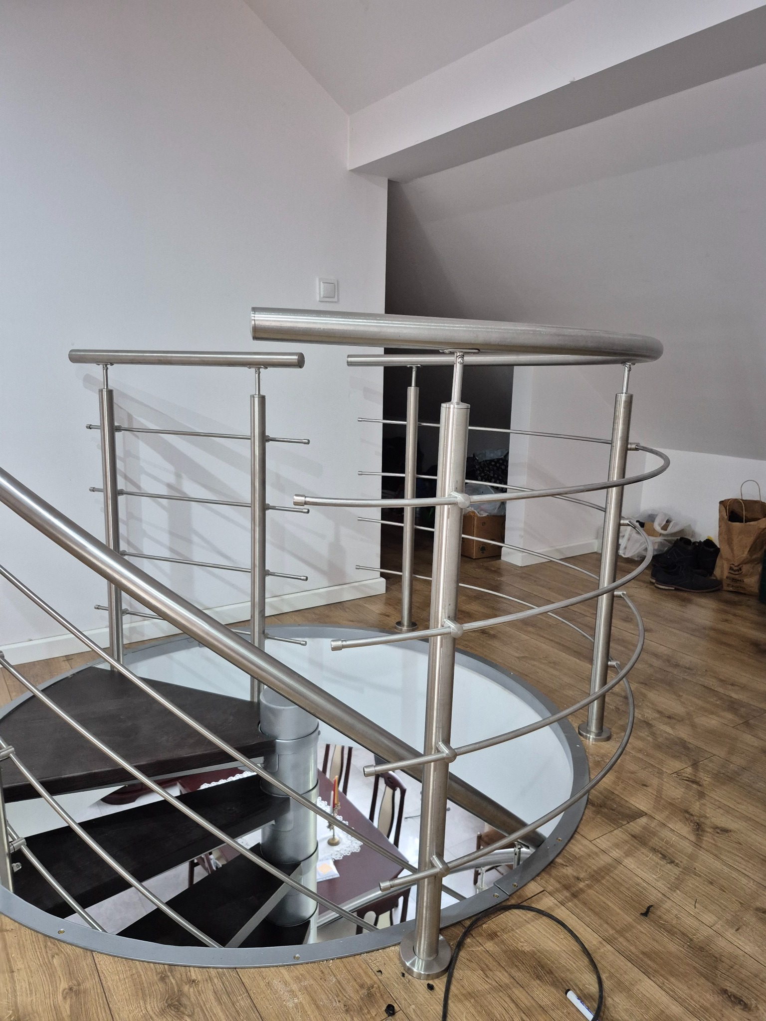 Spiralne schody z balustradą ze stali nierdzewnej, widok z góry, drewniana podłoga, białe ściany. Nietypowy kształt schodów.