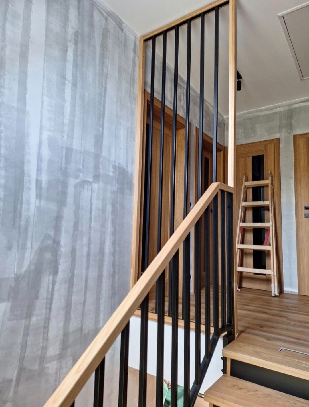 Nowoczesna balustrada schodowa z czarnymi, pionowymi prętami i jasnym drewnianym obramowaniem, na tle ściany o fakturze betonu i drewnianych drzwi.