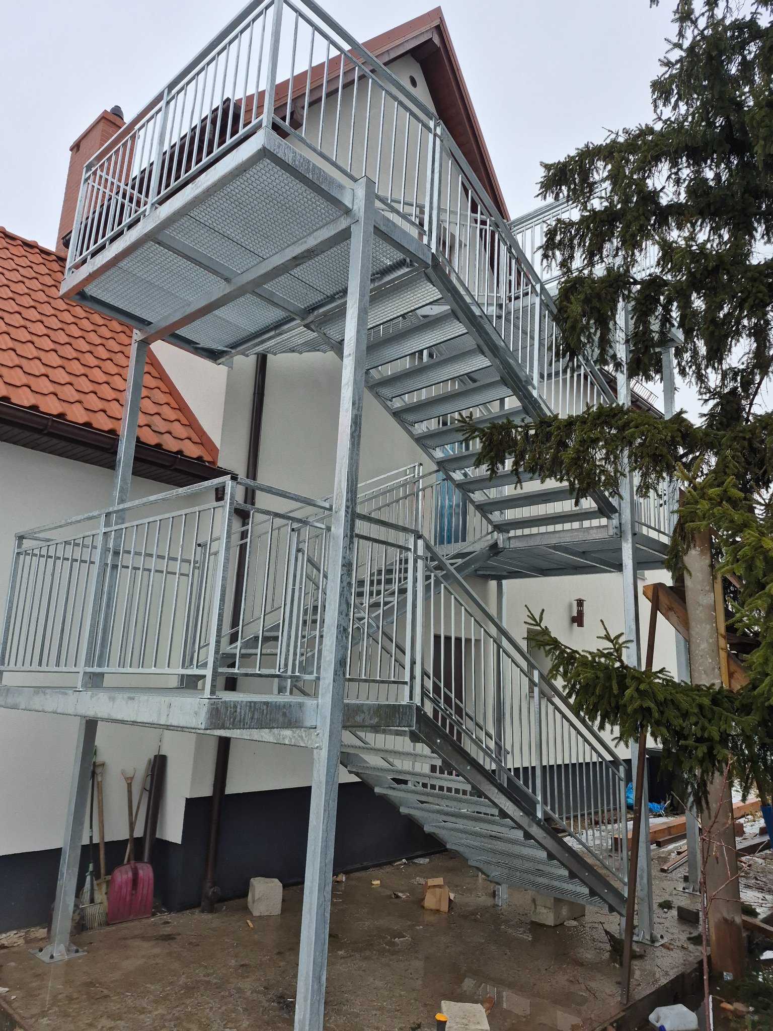 Zewnętrzne schody metalowe z balustradą, prowadzące na balkon. Konstrukcja ocynkowana, ażurowa podłoga, widok z dołu na tle domu z czerwoną dachówką i zielonego drzewa.