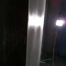 Fragment ściany z nałożoną świeżą warstwą szpachli, oświetlony lampą halogenową na trójnogu, widoczna surowa powierzchnia ściany obok.