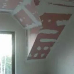 Sufit i skośna ściana poddasza z płyt kartonowo-gipsowych, wstępnie obrobione łączenia i wkręty, widoczne ślady szpachli w kolorze r&oacute;żowym, okno w tle.