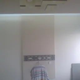 Ściana z zamontowanym panelem i uchwytem TV, na drabinie leży tkanina w paski. Widoczna lampa sufitowa. Neutralna kolorystyka wnętrza.