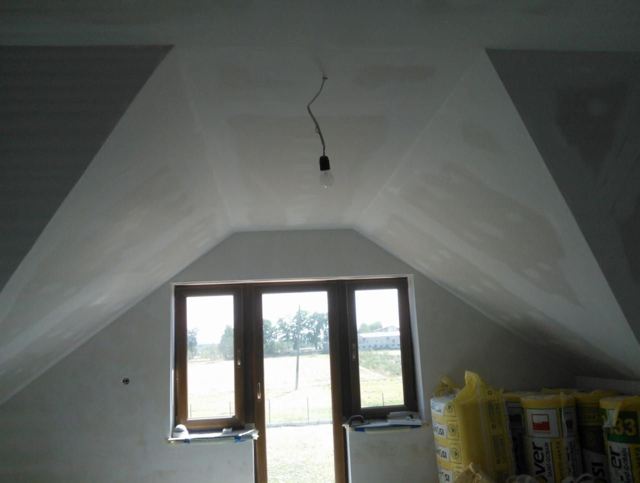 Poddasze z oknem po szpachlowaniu, widoczne nierówności na skośnych ścianach i suficie, przygotowane do dalszych prac wykończeniowych, izolacja przy ścianie.