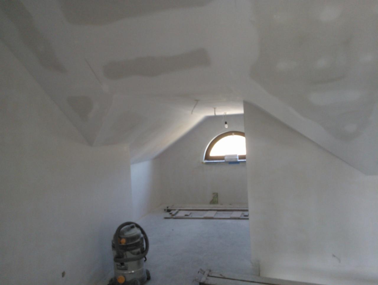 Poddasze po szpachlowaniu, przygotowane do dalszych prac wykończeniowych. Widoczne ślady po łatach na ścianach i suficie, okrągłe okno w tle, odkurzacz przemysłowy.