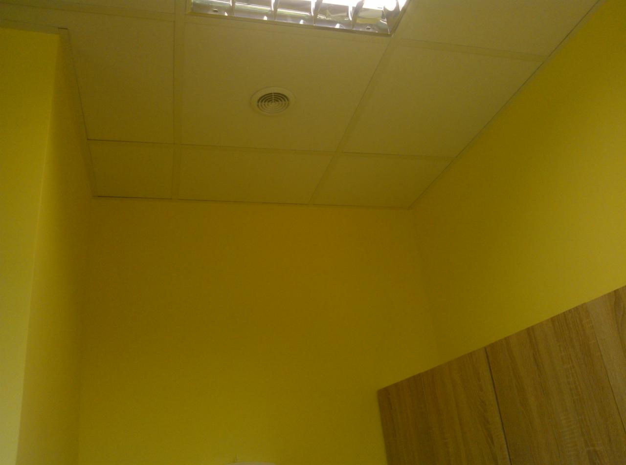 Żółta ściana i sufit podwieszany z kratką oświetleniową i kratką wentylacyjną. Fragment szafki z forniru drewnopodobnego w prawym dolnym rogu.