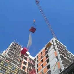 Wysokobudżetowa budowa wielopiętrowego budynku z cegły, z rusztowaniami i dźwigiem podnoszącym pojemnik z zaprawą do tynkowania, widok z dołu na tle błękitnego nieba.