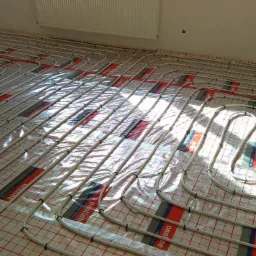 Białe rury ogrzewania podłogowego ułożone na folii z czerwoną siatką. Widoczne mocowania rur. Grzejnik na ścianie w tle. Jasne światło słoneczne.