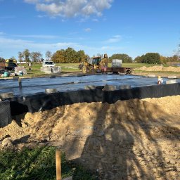 Marcin Raczewski M&M Usługi Remontowo-Budowlane - Fundament domu jednorodzinnego w trakcie budowy, widoczne bloczki fundamentowe i folia hydroizolacyjna, na tle krajobrazu z drzewami i błękitnym niebem.