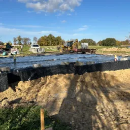 Fundament domu jednorodzinnego w trakcie budowy, widoczne bloczki fundamentowe i folia hydroizolacyjna, na tle krajobrazu z drzewami i błękitnym niebem.