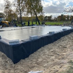 Marcin Raczewski M&M Usługi Remontowo-Budowlane - Fundament domu w trakcie budowy: świeżo wylana płyta, izolacja pionowa, bloczki betonowe. W tle koparka i pracownicy, jesienne drzewa, błękitne niebo z chmurami.