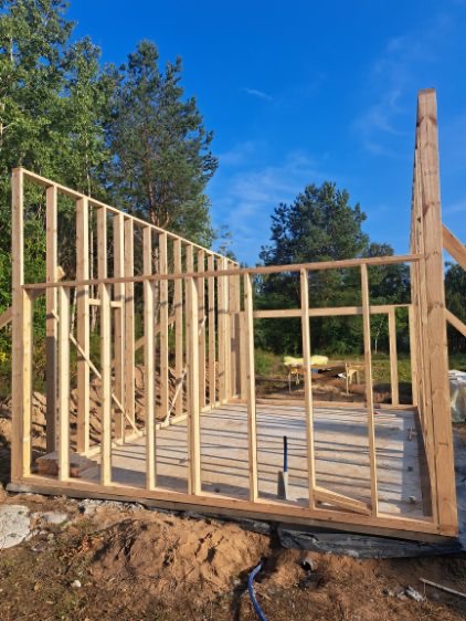 Wznoszenie szkieletu drewnianego budynku na fundamencie, widok z zewnątrz w słoneczny dzień. W tle drzewa i błękitne niebo. Widoczny fundament i częściowo wzniesione ściany.