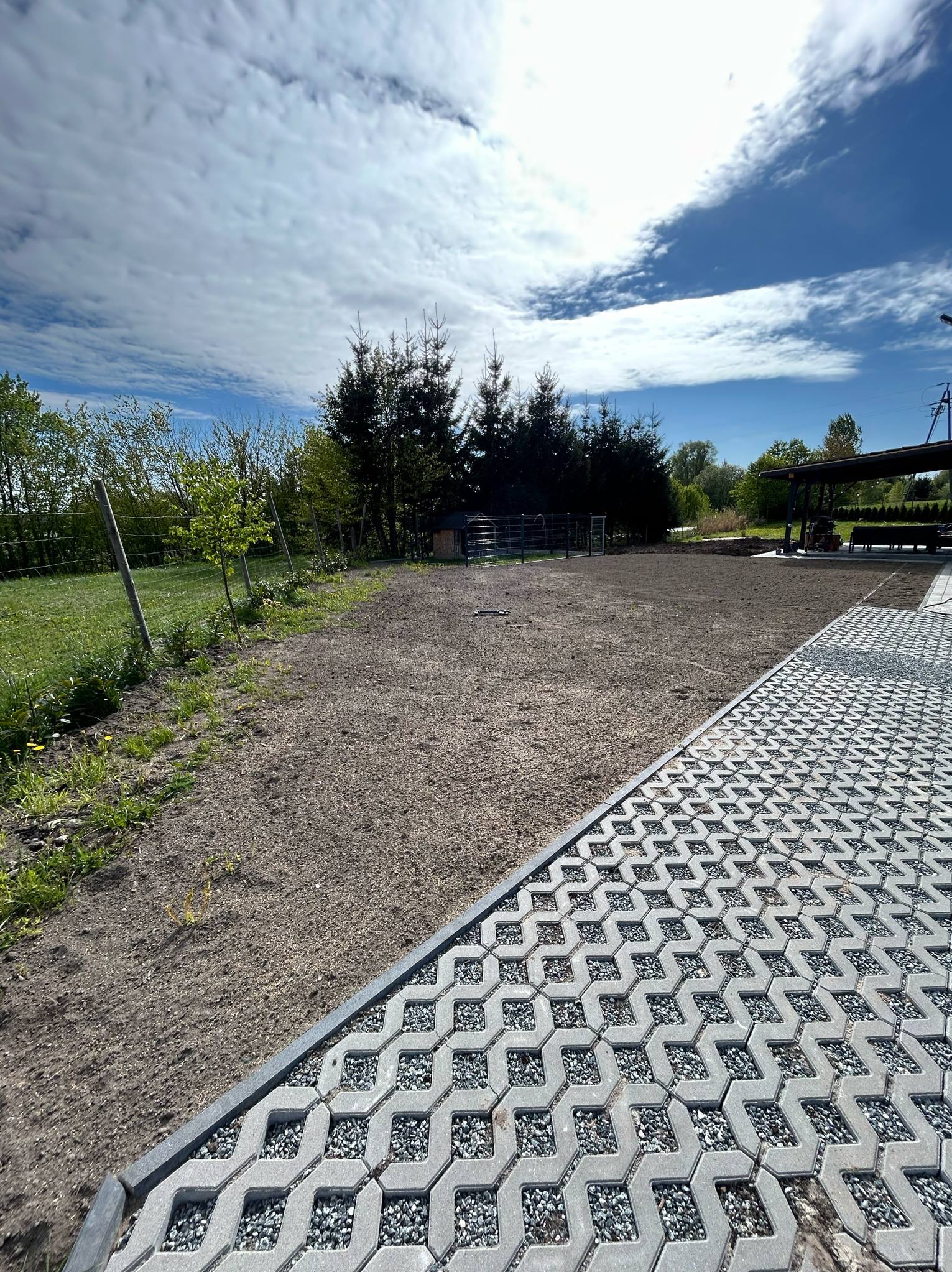 Ułożona szara kostka ażurowa z kamyczkami, widok na przygotowany teren pod trawnik, w tle zieleń drzew i błękitne niebo z chmurami.
