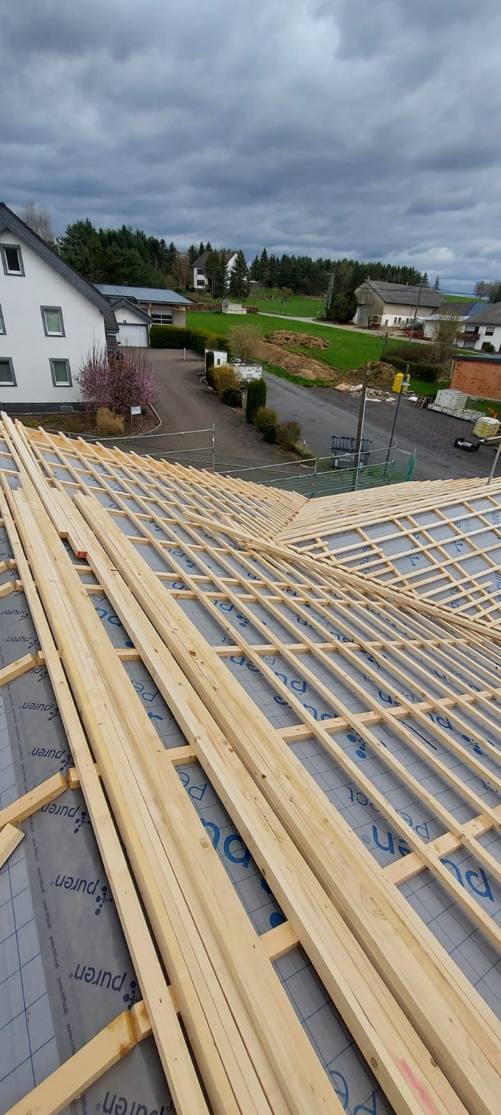 Konstrukcja dachu z drewnianych łat i kontrłat, zabezpieczona membraną, w tle zabudowa wiejska i pochmurne niebo. Widok z góry na szkielet dachu.