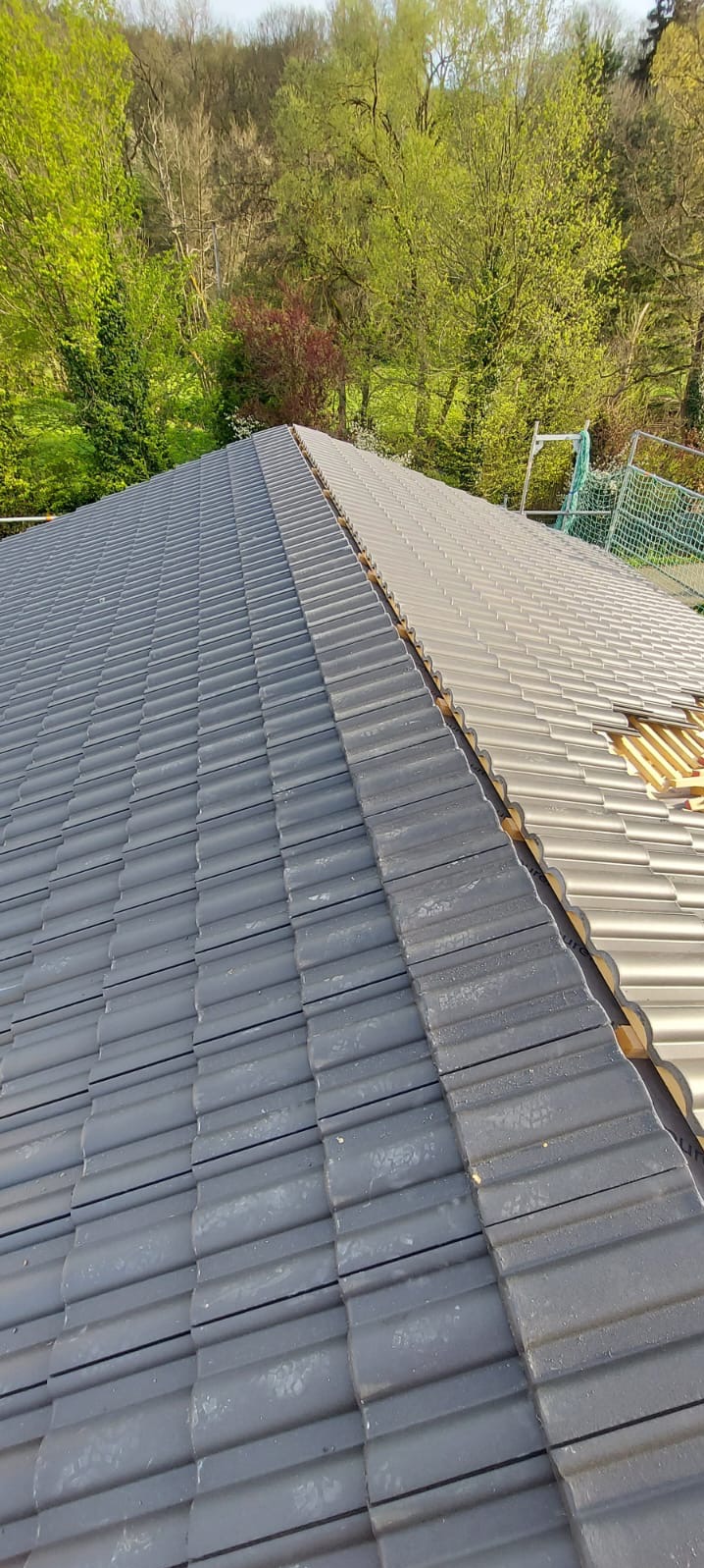 Częściowo wymienione dachówki na dachu domu, widok z góry. Połowa dachu pokryta ciemnoszarą dachówką, druga połowa w trakcie wymiany, widoczne łaty i jasnoszare dachówki.