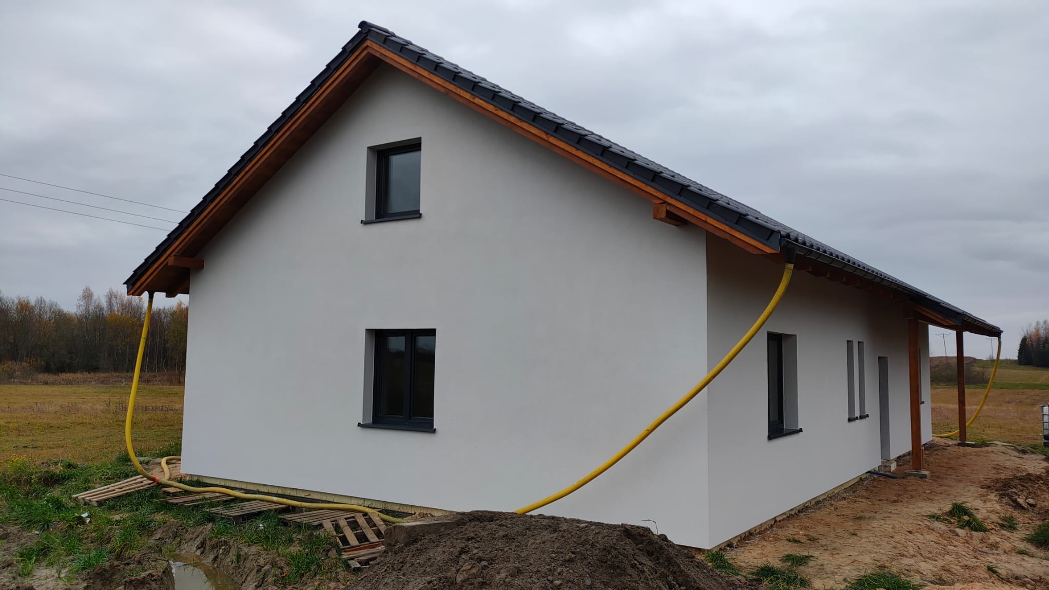 Nowoczesny dom jednorodzinny z szarą elewacją i czarną dachówką, widoczny z boku, z żółtym wężem odprowadzającym wodę deszczową. Tło stanowią drzewa.