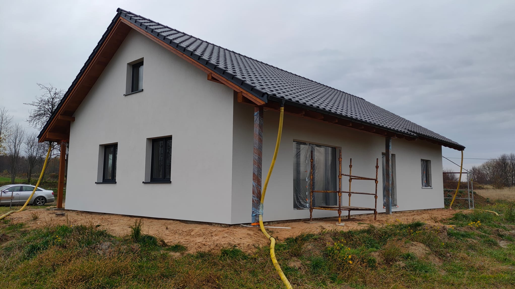 Nowo otynkowany dom jednorodzinny z ciemną dachówką, częściowo osłoniętymi oknami i rusztowaniem. Żółty wąż leży na ziemi. Widok z boku na szarym tle nieba.