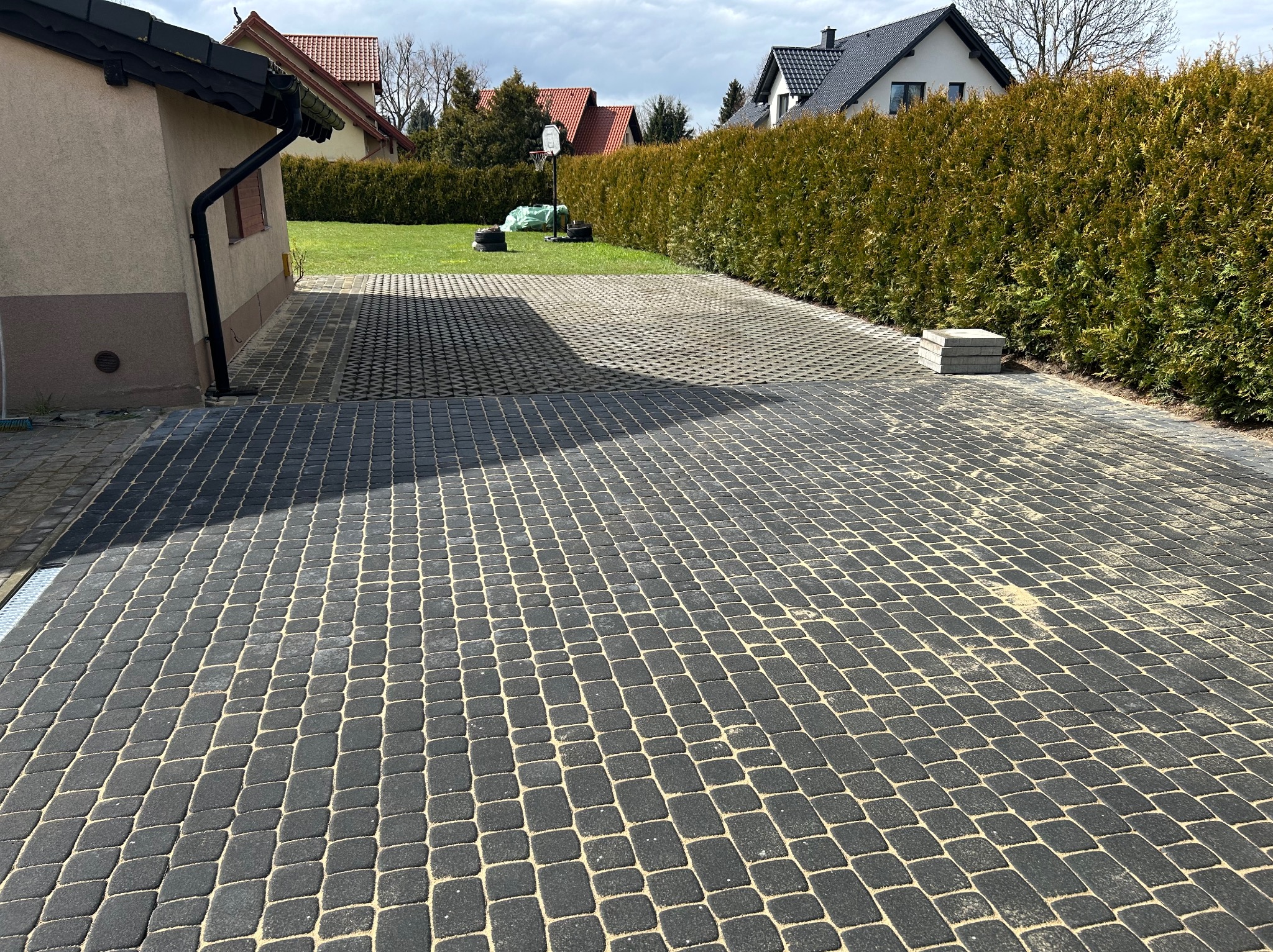 Ułożona, ażurowa nawierzchnia z kostki brukowej w kolorze grafitowym, wypełniona piaskiem, tworząca podjazd do domu z zielonym trawnikiem i tujami w tle.