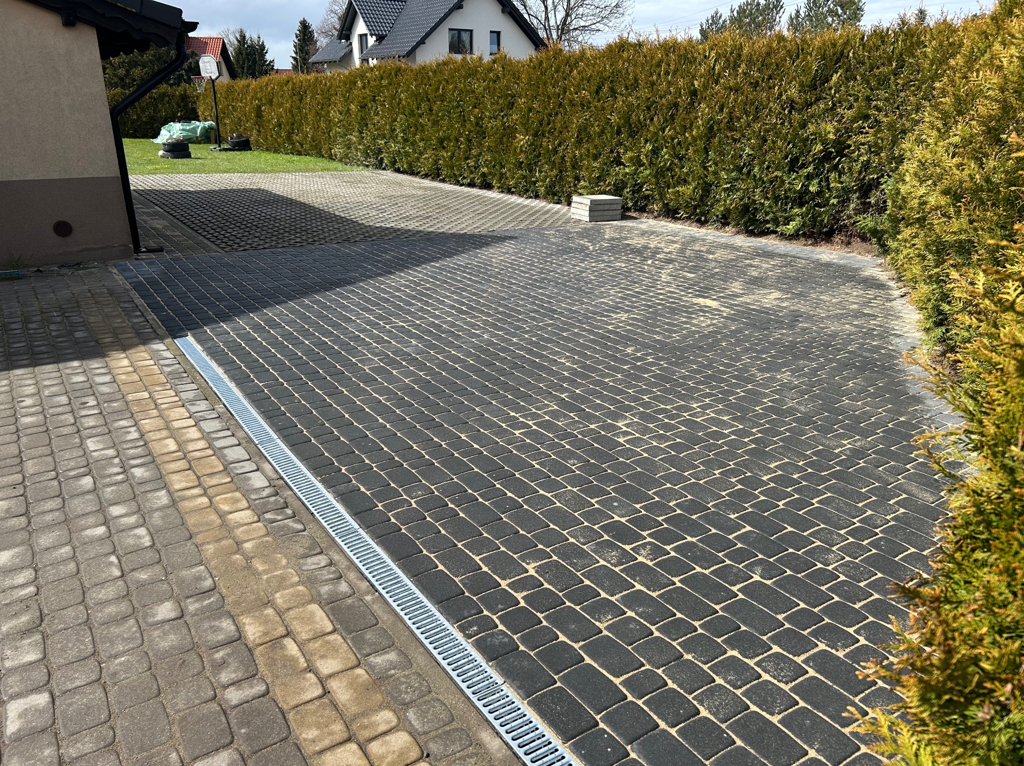 Ułożona kostka brukowa w szarym odcieniu, z widocznym systemem odprowadzania wody. W tle żywopłot z tui i budynek mieszkalny.
