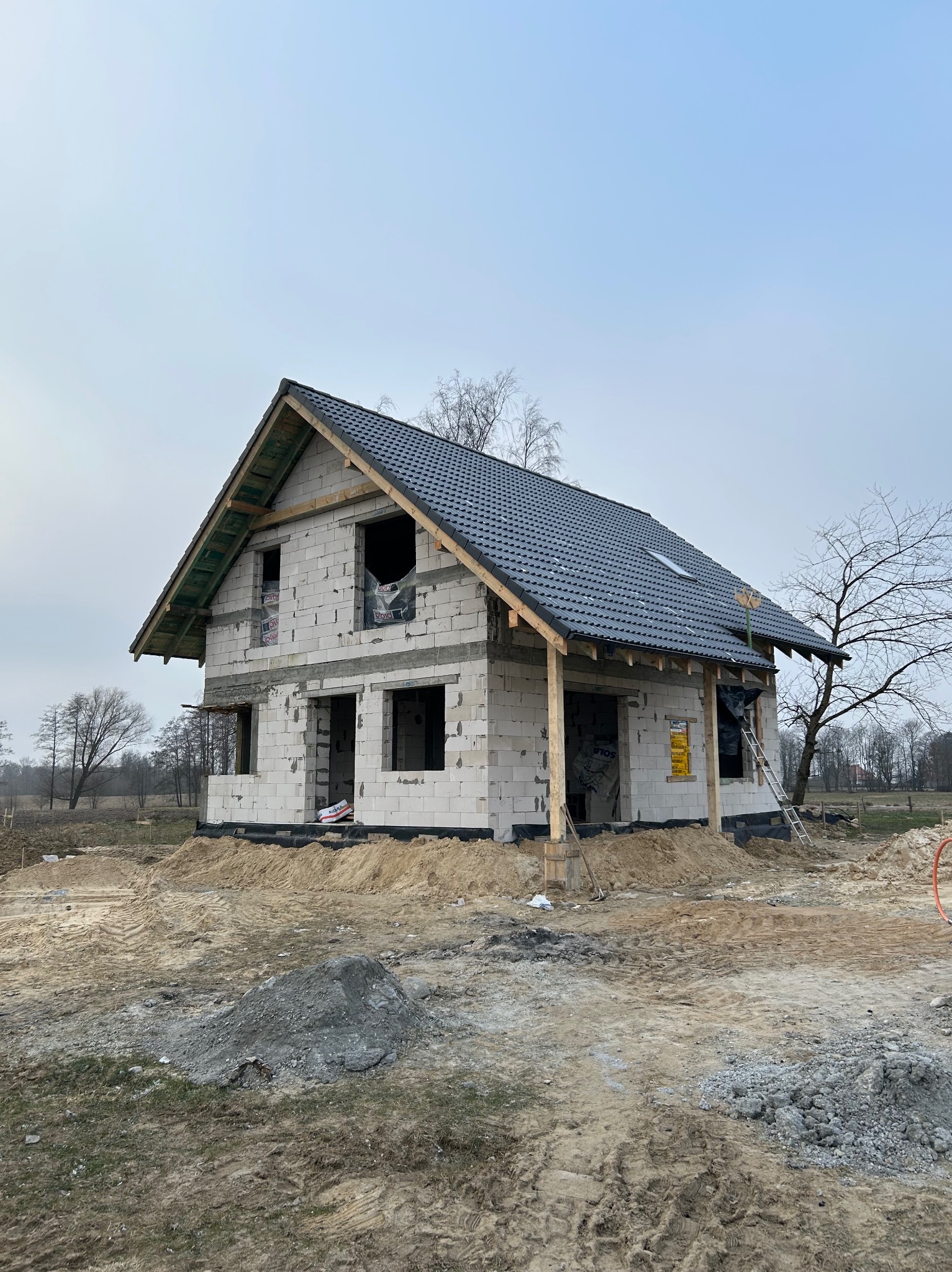 Surowy dom z białego pustaka z ciemną dachówką w trakcie budowy, widok z zewnątrz, na pierwszym planie ziemia i gruz. Niebo pochmurne, drzewa bez liści w tle.