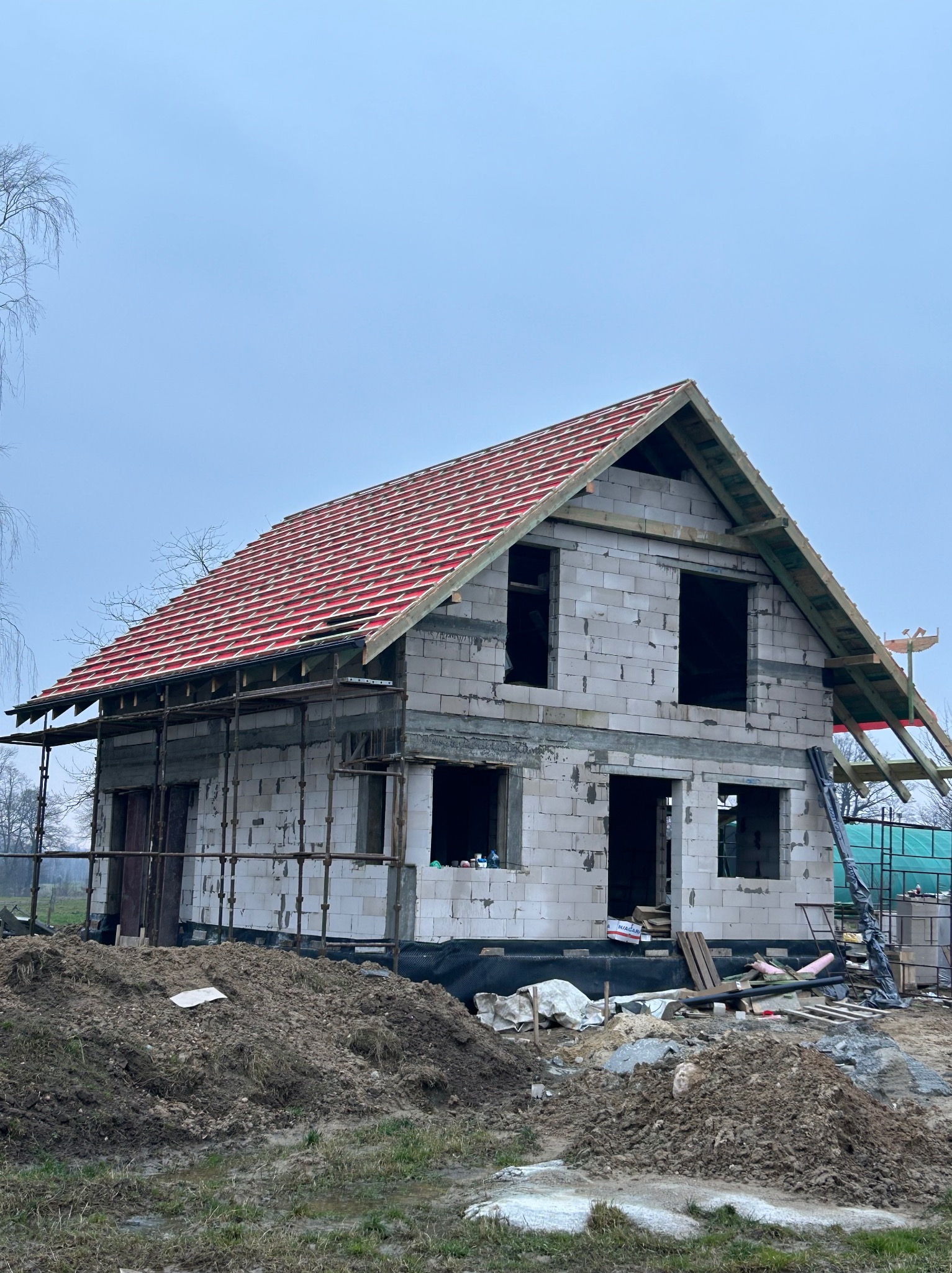 Budowa domu jednorodzinnego w stanie surowym otwartym. Widoczne ściany z bloczków, częściowo ułożona dachówka i rusztowania. Prace budowlane w toku.