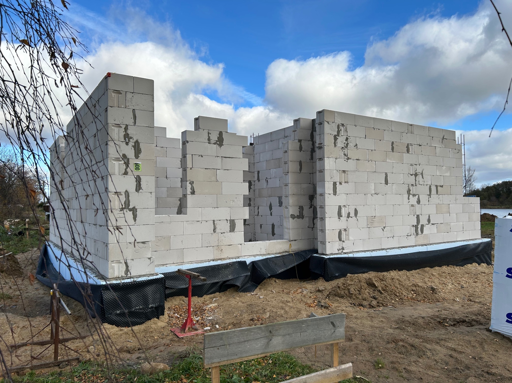 Wznoszenie ścian domu z bloczków betonu komórkowego. Widoczne czarne zabezpieczenie fundamentów. Częściowo zbudowane ściany na tle błękitnego nieba.