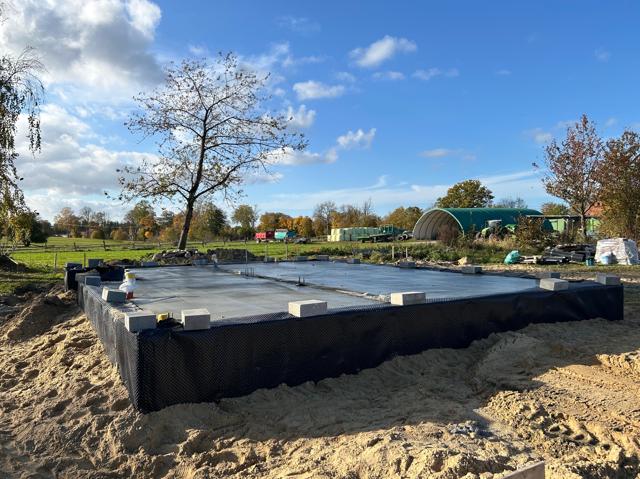 Fundament płyty budynku w trakcie budowy, zabezpieczony czarną folią, z widocznymi elementami konstrukcyjnymi i otaczającym terenem budowy z piaskiem.