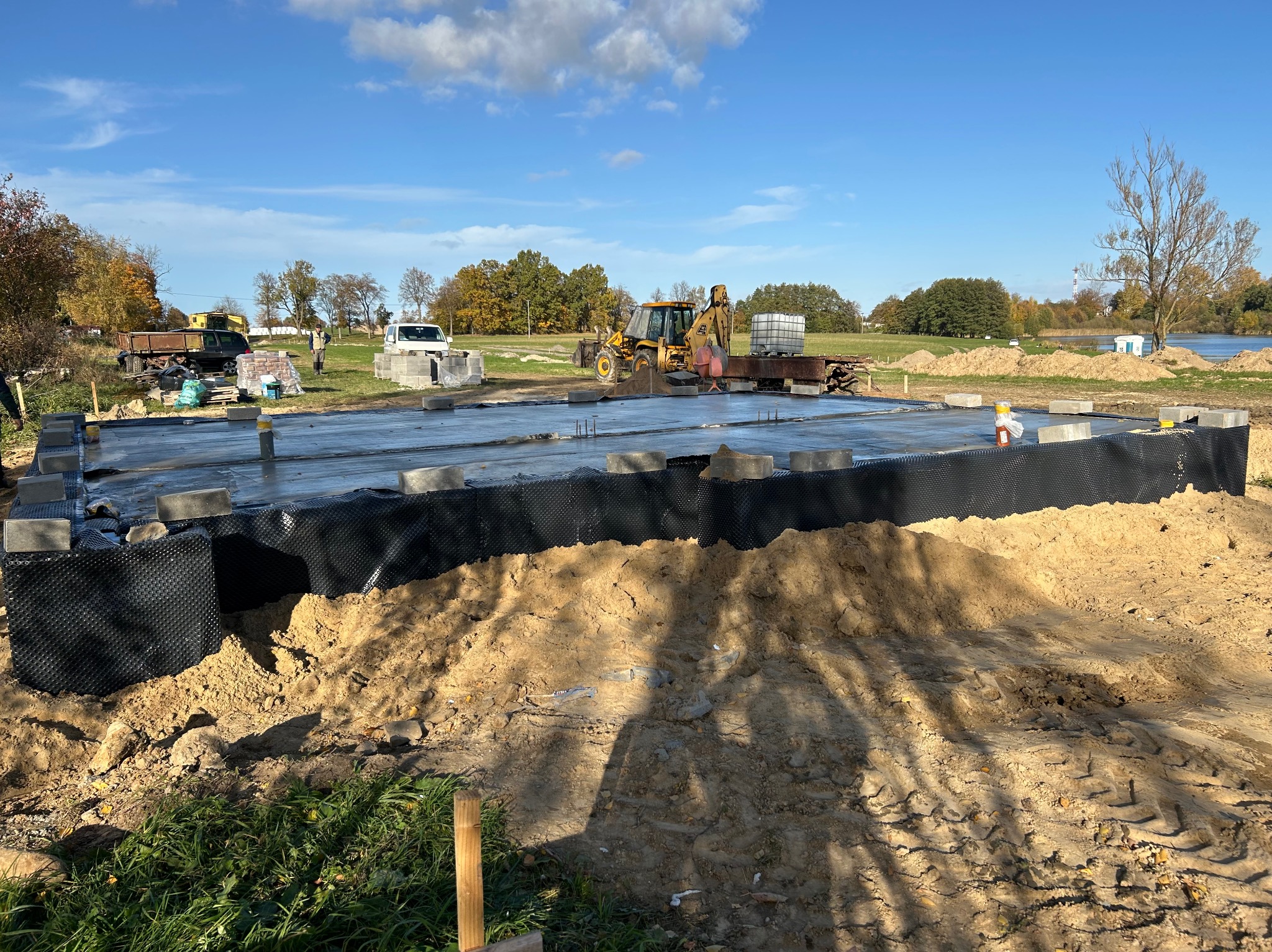 Fundament domu jednorodzinnego w trakcie budowy, widoczne bloczki fundamentowe i folia hydroizolacyjna, na tle krajobrazu z drzewami i błękitnym niebem.