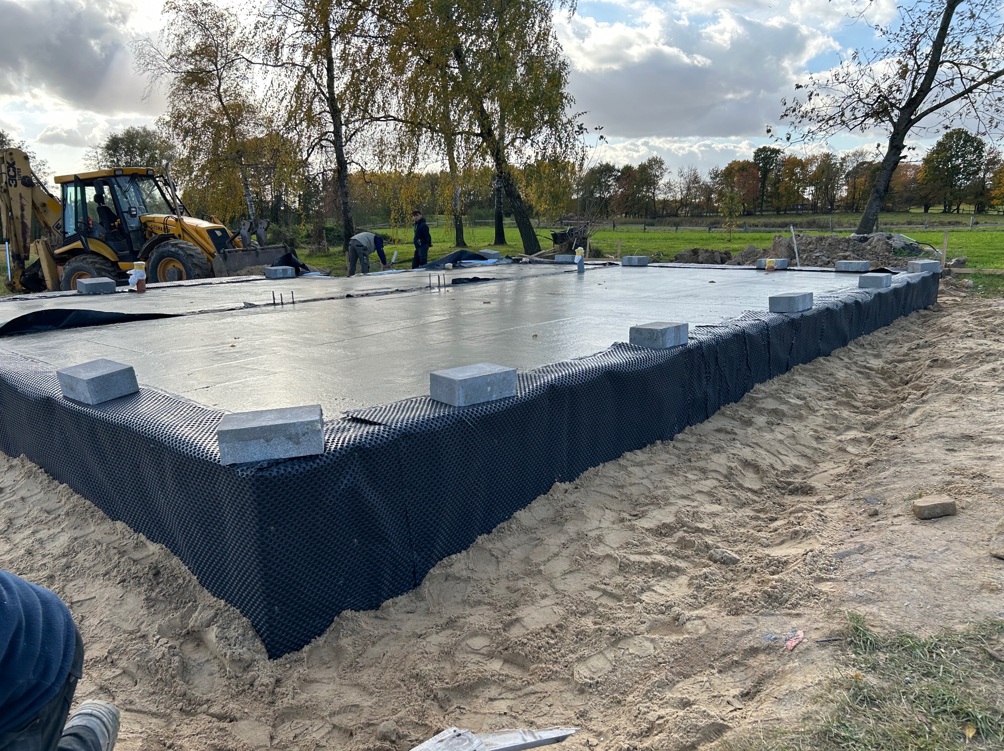 Fundament domu w trakcie budowy: świeżo wylana płyta, izolacja pionowa, bloczki betonowe. W tle koparka i pracownicy, jesienne drzewa, błękitne niebo z chmurami.