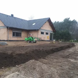 Nowo powstający dom jednorodzinny w stanie surowym otoczony wykopami i stertą żwiru, z widocznym zielonym miniładowarką na działce, w tle las.