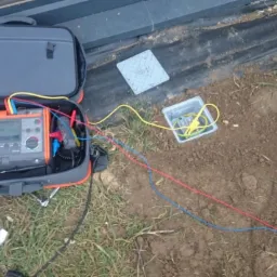 Miernik w walizce, kable podłączone do ziemi w plastikowej skrzynce. Pomiary elektryczne w terenie, narzędzia obok.