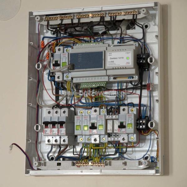 Rozdzielnica elektryczna w obudowie, widoczne bezpieczniki, przewody i moduły sterujące. Kompaktowy układ, detale połączeń.