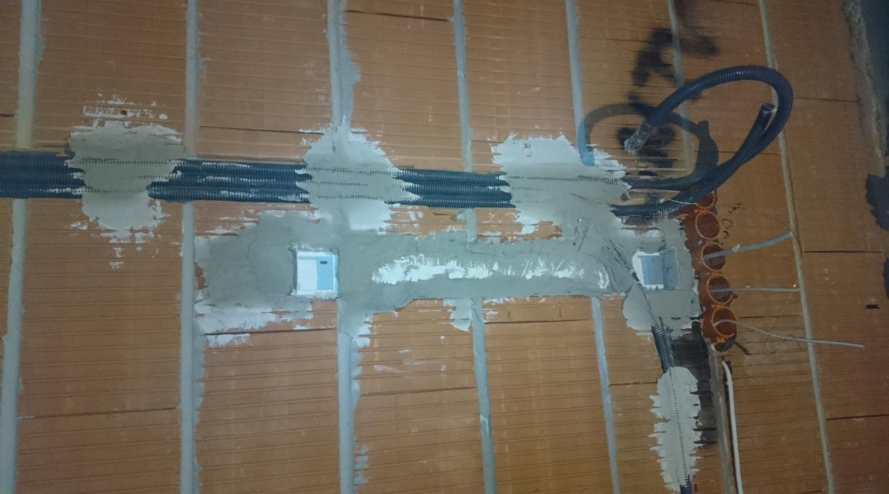 Instalacja przewodów elektrycznych w bruzdach ceglanej ściany, zabezpieczonych siatką i zaprawą. Widoczne puszki instalacyjne i peszle. Prace w toku.