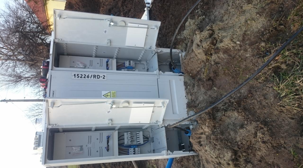 Otwarta, biała skrzynka elektryczna 15226/RD-2 w ziemi, z widocznymi przewodami i elementami instalacji. Na skrzynce naklejka ostrzegawcza.