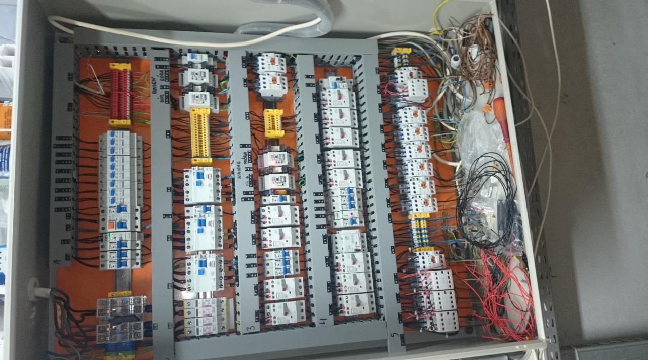 Rozdzielnica elektryczna z licznymi zabezpieczeniami i okablowaniem, widok z góry, wnętrze szafy z elementami instalacji elektrycznej w kolorach: pomarańczowym, żółtym, białym.