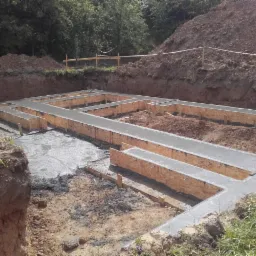 Wylane betonowe fundamenty w drewnianym szalunku, widoczne wystające pręty zbrojeniowe, teren budowy z ziemią i zielenią w tle.