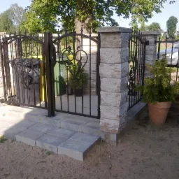 Metalowa brama z kamiennymi słupkami i schodami w Kołobrzegu. Dekoracyjne elementy z motywem serca. Ustawione donice z roślinami. Widoczny fragment ogrodzenia i samochodu.