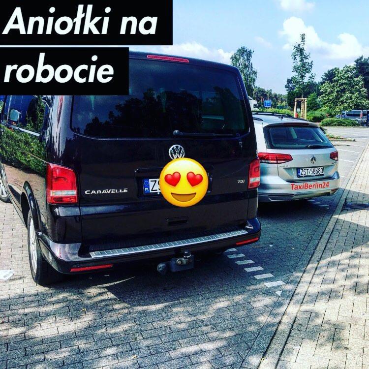 Tył czarnego Volkswagena Caravelle z hakiem holowniczym i naklejką z emotikoną na tablicy rejestracyjnej, obok srebrny samochód z napisem TaxiBerlin24 na parkingu.