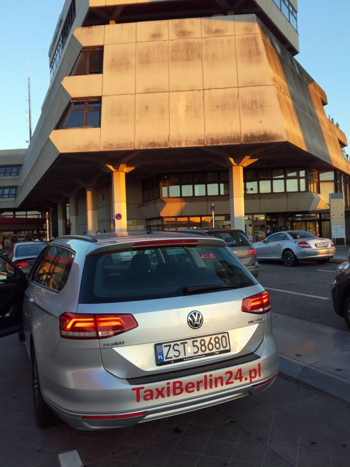 Srebrny Volkswagen Passat kombi z otwartymi drzwiami, oznaczony jako TaxiBerlin24.pl, zaparkowany przed betonowym budynkiem w słoneczny dzień.