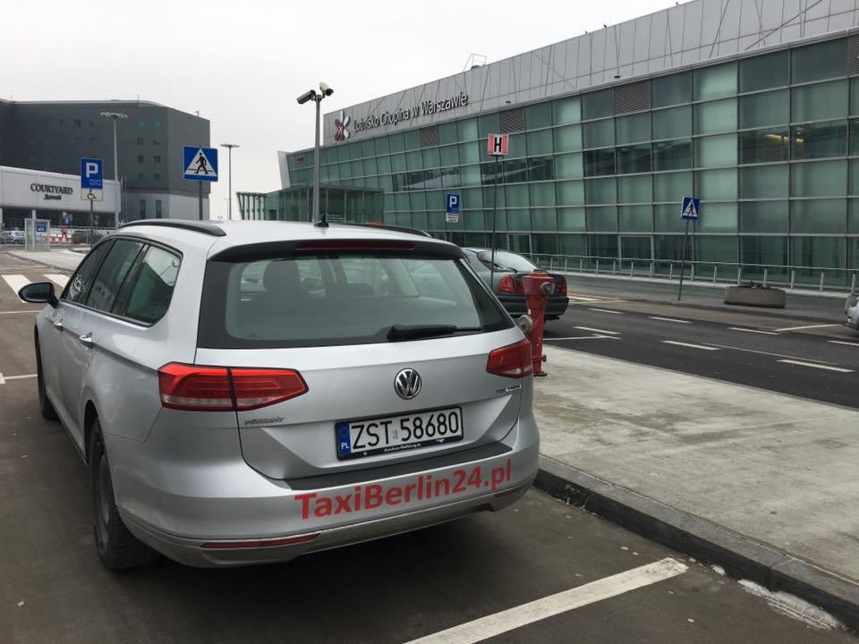 Srebrny Volkswagen Passat kombi z logo TaxiBerlin24.pl zaparkowany przed lotniskiem Chopina w Warszawie, widok z tyłu.