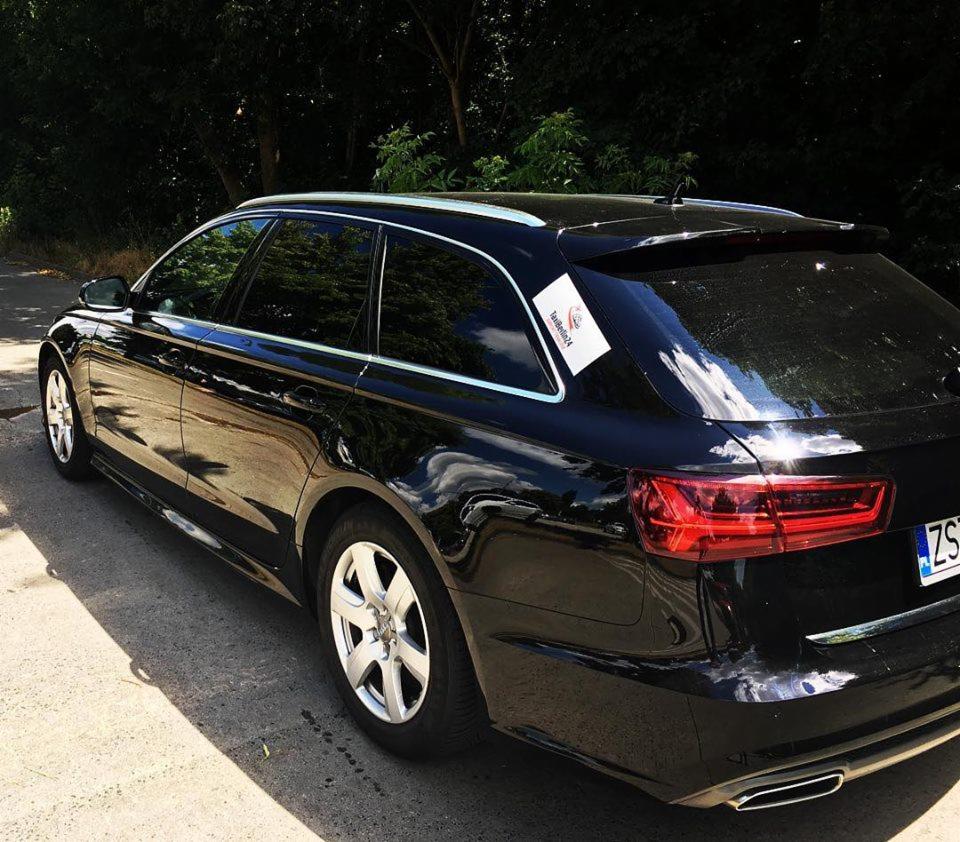 Czarny samochód typu kombi z naklejką TaxiBerlin24 na tylnej szybie, zaparkowany na ulicy w słoneczny dzień.