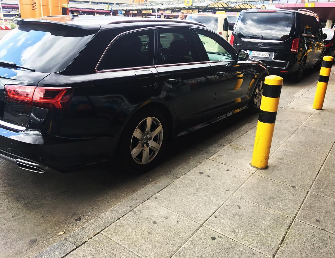 Czarny samochód kombi Audi A6 zaparkowany obok czarnego vana Mercedes V250d z niemieckimi tablicami rejestracyjnymi i żółto-czarnymi słupkami ostrzegawczymi na chodniku.