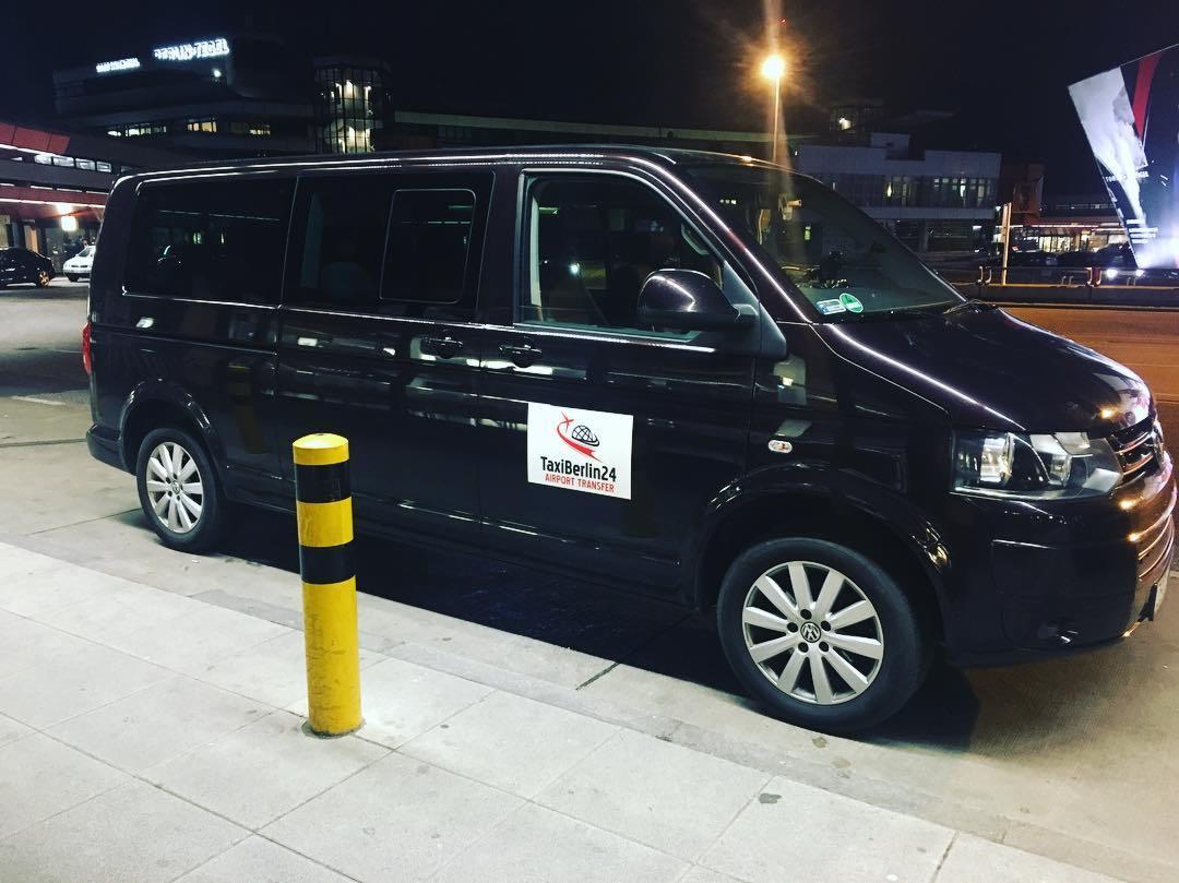 Czarny van z logo TaxiBerlin24 Airport Transfer zaparkowany przy żółto-czarnym słupku na tle budynku lotniska nocą.