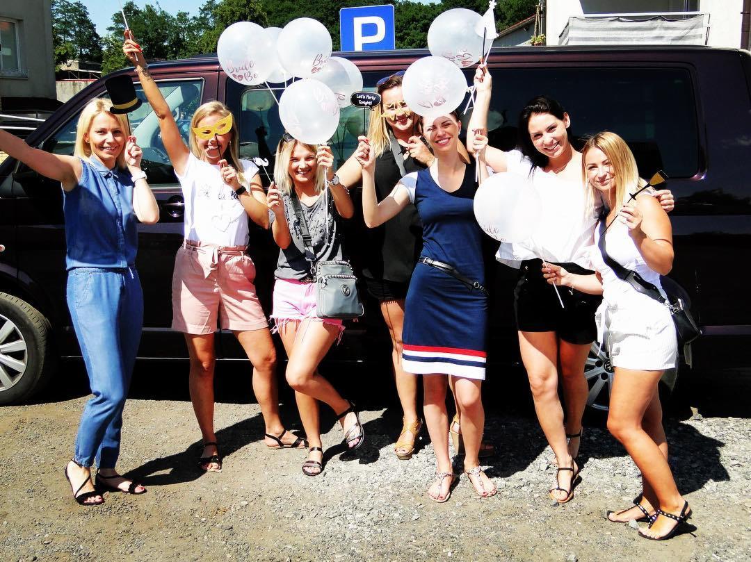 Grupa siedmiu uśmiechniętych kobiet świętuje przed ciemnym vanem, trzymając białe balony z napisem 'Bride to be' i rekwizyty do zdjęć.