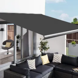 Rozwinięta, grafitowa osłona przeciwsłoneczna nad tarasem z meblami ogrodowymi, widok na wnętrze domu przez przesuwne drzwi, na zewnątrz drewniany płot i zieleń.